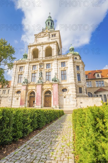 Imposing building with towers, green roofs and a cobbled path surrounded by a garden, Munich, Germany