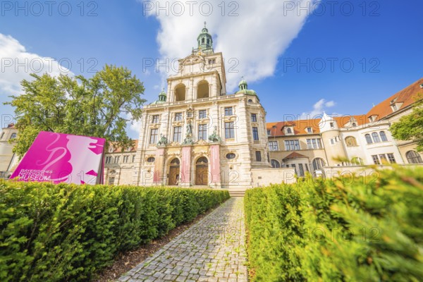 Historic building with towers and pink banners framed by manicured hedges and blue sky, Munich, Germany