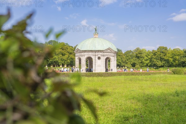 Meadow environment around grey pavilion with green dome, clothed by resting people and blue sky, Munich, Germany