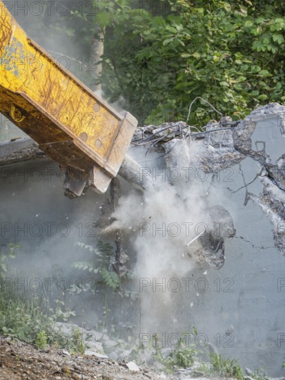 An excavator tears down a concrete structure with its shovel, surrounded by forest, dusty atmosphere, bridge demolition, Calw, Germany