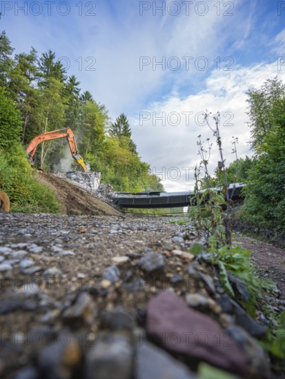 Wide angle shot of an excavator on a gravel road demolishing a bridge, surrounded by lush greenery and cloudy sky, bridge demolition, Calw, Germany