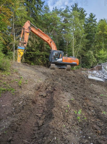 An excavator working on a pile of earth in a wooded area with a clear sky, bridge demolition, Calw, Germany