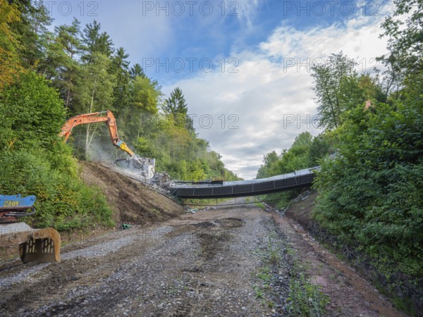 Two excavators tearing down a bridge on a gravel road, flanked by forest and a cloudy sky, bridge demolition, Calw, Germany