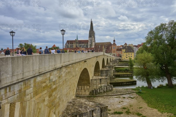 People walking across a historic bridge with a view of the cathedral and trees, Stone Bridge, Danube, Regensburg, Bavaria, Germany