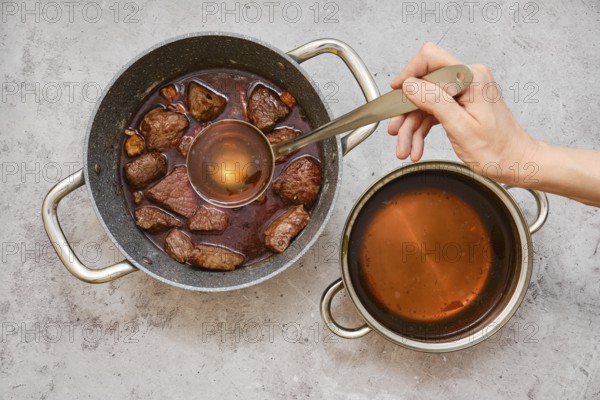 A person is using a ladle to add beef broth to a pot filled with fried chunks of beef shoulder and vegetables