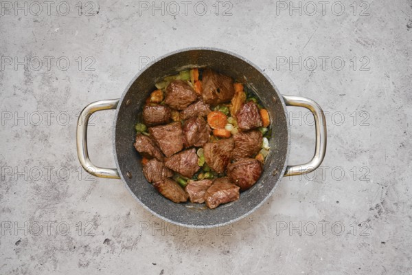 Tender pieces of beef are simmering with a mix of carrots, celery, and onions in a cooking pot. The meal is being prepared on a neutral-toned countertop in a contemporary kitchen setting