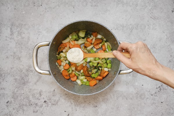Freshly chopped carrots, celery, and onions are fried in a pot. A hand is sprinkling flour over the colourful mixture, indicating the preparation of a savory dish in a home kitchen