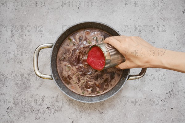 A hand pours diced tomatoes from a can into a pot filled with a rich, simmering beef stew