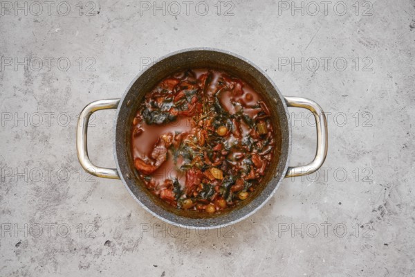 A large pot filled with a flavourful beef and vegetable stew on a gray kitchen countertop. Ingredients like tomatoes and leafy greens are visible, suggesting a healthy home-cooked meal