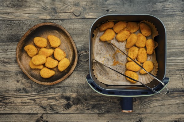 Golden and crispy chicken nuggets rest in an air fryer beside a wooden plate filled with freshly cooked pieces. The meal is prepared on a rustic wooden table, perfect for sharing