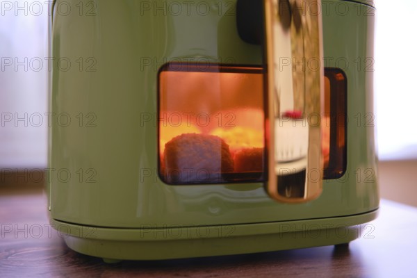 Golden-brown chicken nuggets are cooking in an air fryer, with a warm glow visible through the glass window. The cozy atmosphere suggests a leisurely afternoon meal preparation at home