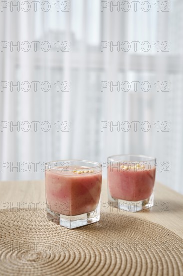 Two elegant glasses filled with a vibrant banana-strawberry smoothie sit atop a woven placemat on a wooden table. Soft natural light filters through sheer curtains, enhancing the inviting atmosphere