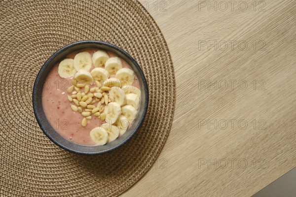 Bright and colorful banana strawberry smoothie is topped with fresh banana slices and pine nuts, presented in a bowl placed on a woven placemat on a wooden tabletop, perfect for a healthy snack