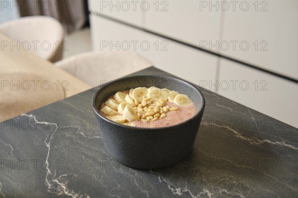 Bright and inviting banana-strawberry smoothie sits in a dark bowl, garnished with fresh banana slices and pine nuts. The cozy kitchen provides a warm backdrop for this healthy treat