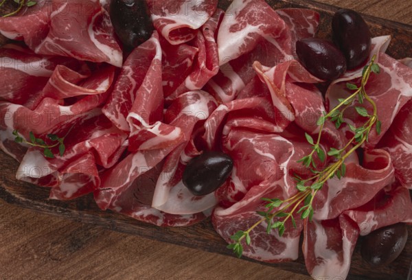 Freshly cut dried meat, served with olives and herbs, on a wooden chopping board, no people