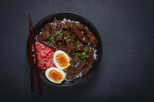 Udon Japanese beef with rice, pickled ginger, boiled egg slice, garnished with green onions, in a black bowl, with chopsticks