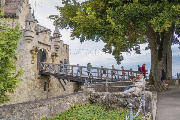 People crossing an old stone bridge towards a castle, surrounded by trees, Lichtenstein Castle, Lichtenstein, Swabian Alb, Germany