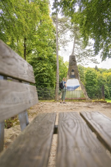 Woman with rucksack looking at a monument in the forest, near a wooden bench, Lichtenstein Castle, Lichtenstein, Swabian Alb, Germany