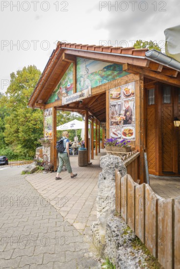 A wooden restaurant at the edge of the forest with passing hikers on a paved path, Lichtenstein Castle, Lichtenstein, Swabian Alb, Germany