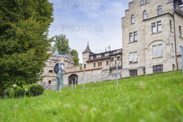 A woman next to a historic building with a green meadow and trees in the background, Lichtenstein Castle, Lichtenstein, Swabian Alb, Germany