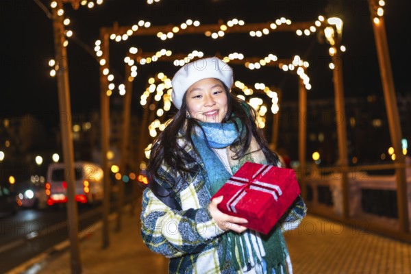 Smiling young woman holding christmas present enjoying winter holidays season under decorative string lights in city street at night
