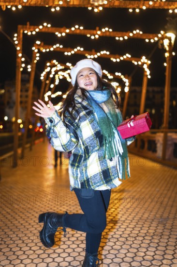 Happy young woman holding a christmas gift, reveling in the enchanting glow of festive lights and decorations on a winter night