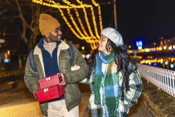 Young multiethnic couple walking arm in arm at illuminated christmas market, man carrying christmas gifts