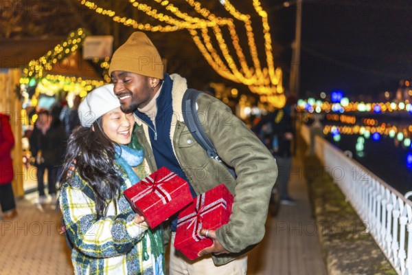 Multiethnic couple embracing and joyfully exchanging christmas gifts at a vibrant christmas market, surrounded by twinkling lights and festive cheer