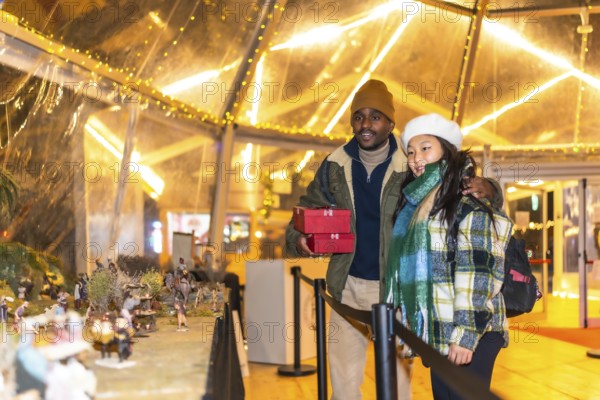 Multiethnic couple holding christmas gifts looking at a nativity scene in a christmas village illuminated at night