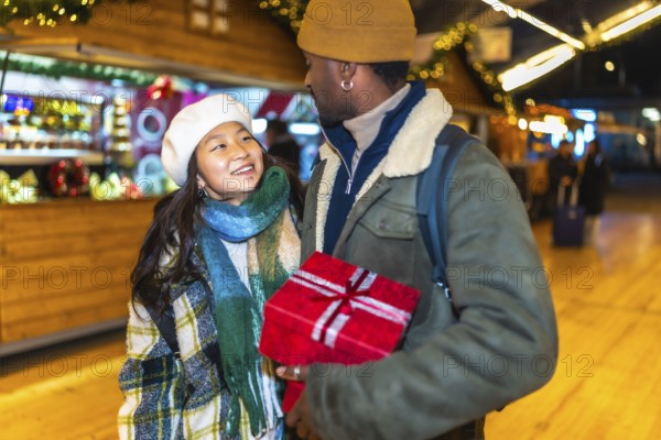 Young multiethnic couple joyfully holding a beautifully wrapped christmas gift while enjoying the festive atmosphere of a decorated market