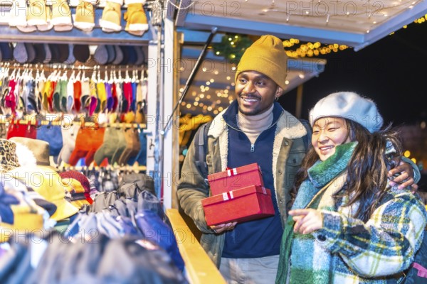 Young multiethnic couple enjoying christmas shopping at a decorated christmas market, holding red gift boxes