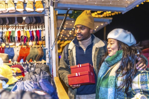 Young multiethnic couple enjoying christmas shopping at a decorated christmas market, holding red gift boxes and smiling