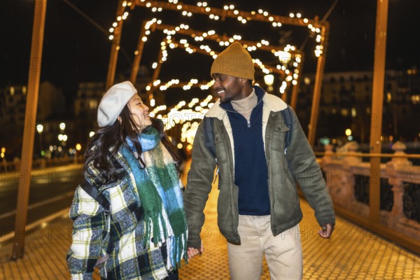 Young multiethnic tourists couple holding hands, enjoying christmas lights decoration at night while walking on a bridge