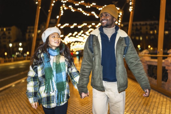 Young multiethnic couple holding hands and walking under christmas lights decorations, enjoying winter holidays together