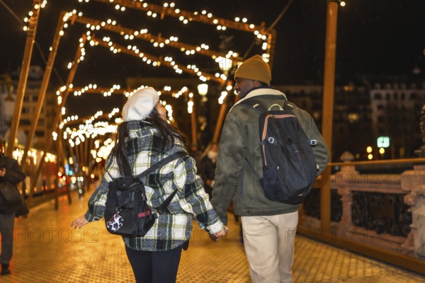 Young multiethnic couple walking on bridge with christmas lights, enjoying winter holidays traveling in europe