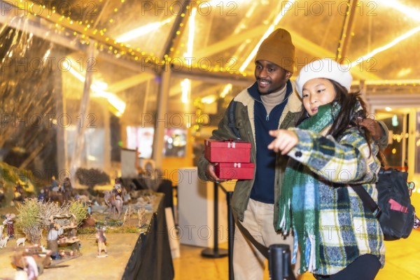 Happy multiethnic couple holding christmas gifts and admiring a nativity scene at a christmas market