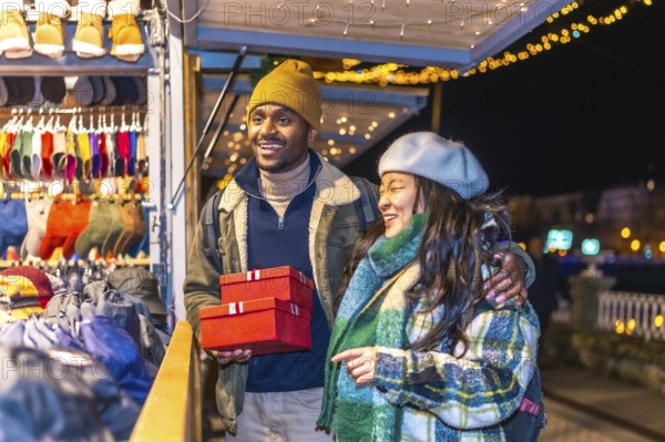 Young multiethnic couple enjoying christmas shopping at a decorated christmas market, holding red gift boxes