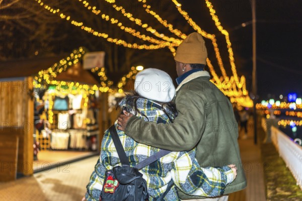Young multiethnic couple walking hand in hand, sharing warm hugs amidst the glowing lights of a festive christmas market at night