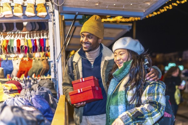 Multiethnic couple enjoying christmas shopping at a decorated christmas market, holding red gift boxes and smiling