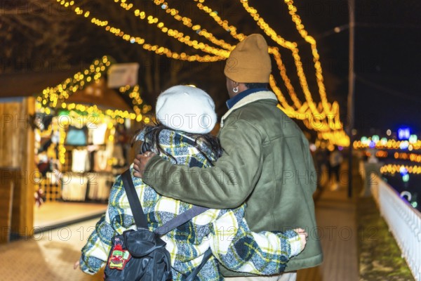 Multiethnic couple strolling through a beautifully decorated outdoor christmas market, embracing the festive spirit of the holiday night