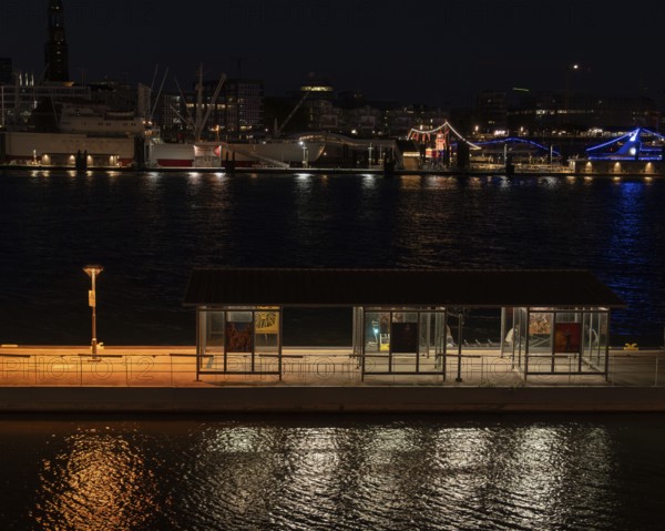 Night shot of a pontoon with bus shelter at the theatre on the Elbe with a view of the lighting of the Landungsbrücken, Hamburg, Germany