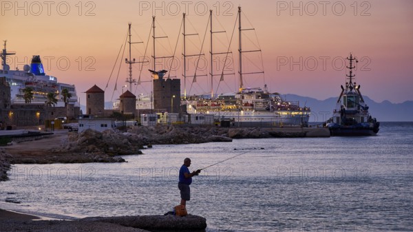 A fisherman fishing at sunset at an illuminated harbour with several ships on the horizon, Rhodes Old Town, Rhodes Town, Rhodes, Dodecanese, Greek Islands, Greece