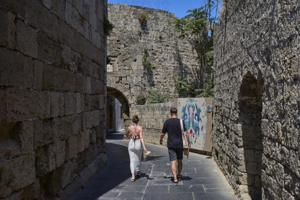 Saint John's Tor, Narrow alleyway lined with old walls with wall art and walkers in sunny weather, Rhodes Old Town, Rhodes Town, Rhodes, Dodecanese, Greek Islands, Greece