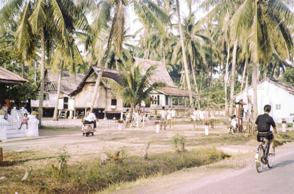 Traditional village houses in coconut palm trees, boys and young men playing Sepak Takraw foot volleyball game, Malacca or Melaka, Malaya, Malaysia, south east Asia c 1964