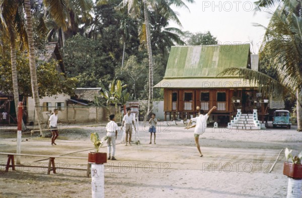 Traditional village houses in coconut palm trees, boys and young men playing Sepak Takraw foot volleyball game, Malacca or Melaka, Malaya, Malaysia, south east Asia c 1964