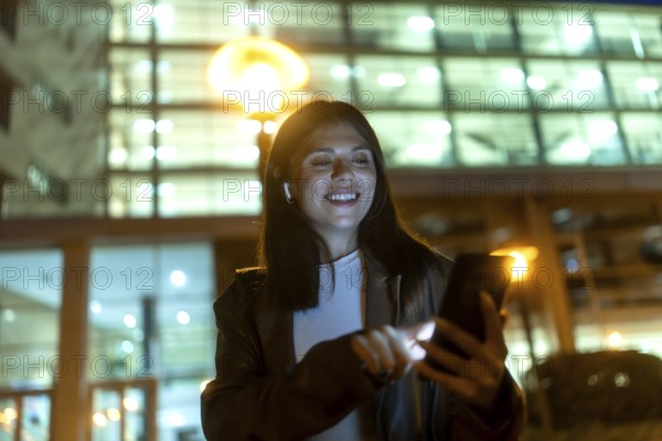 Young woman smiling and engaging with her smartphone, wearing wireless earbuds for connectivity and entertainment while standing in a vibrant city at night