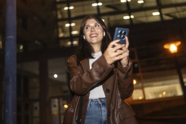 Young woman happily smiling and looking up while using her smartphone and wireless earbuds at night, capturing urban moments or communicating with friends in a city environment