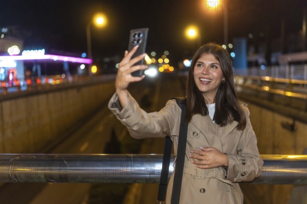 Young woman smiling and taking a selfie with a smartphone at night, capturing moments of connection and urban life on a bridge with city lights in the background