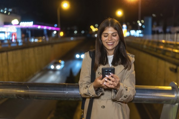 Young woman smiling while looking at her smartphone, standing on an urban bridge with city lights and passing cars creating a blurred background at night