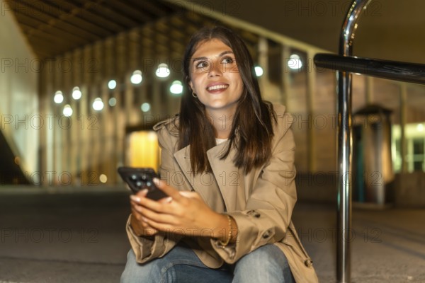 Young woman smiling while holding a mobile phone, looking up and away, sitting in an urban environment at night with bokeh lights, expressing connectivity and waiting